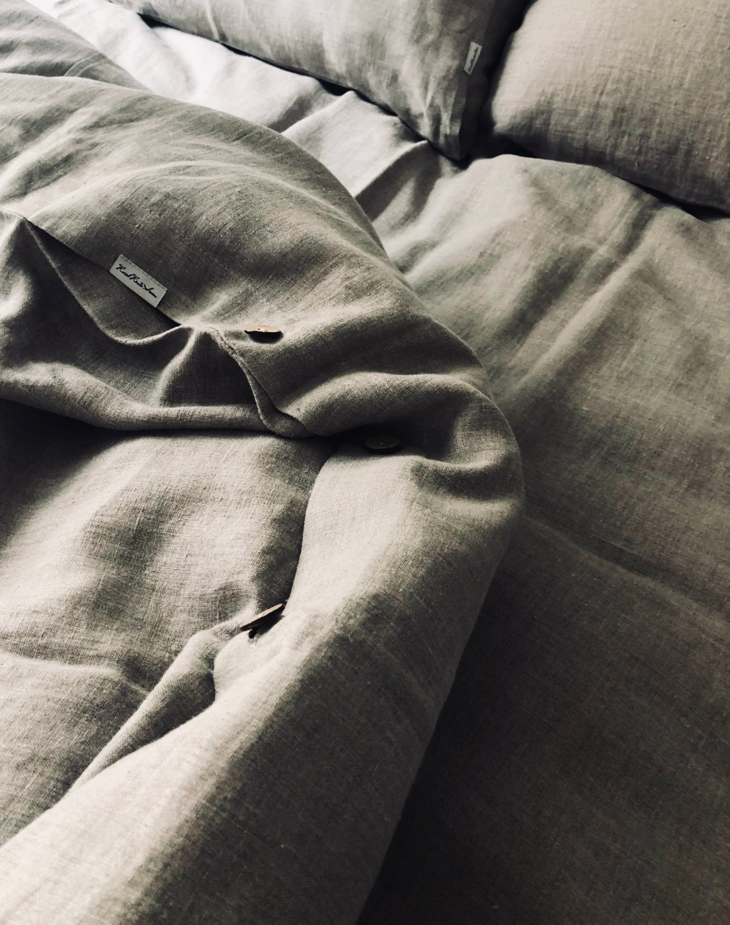Moody photo of turned down bed dressed in Oeko-Tex Certified Organic European Linen Queen size Duvet Set wit two pillowcases showing Rural Route Seven logo brand label and coconut wood buttons with dim moody sunlight streaming in from the window