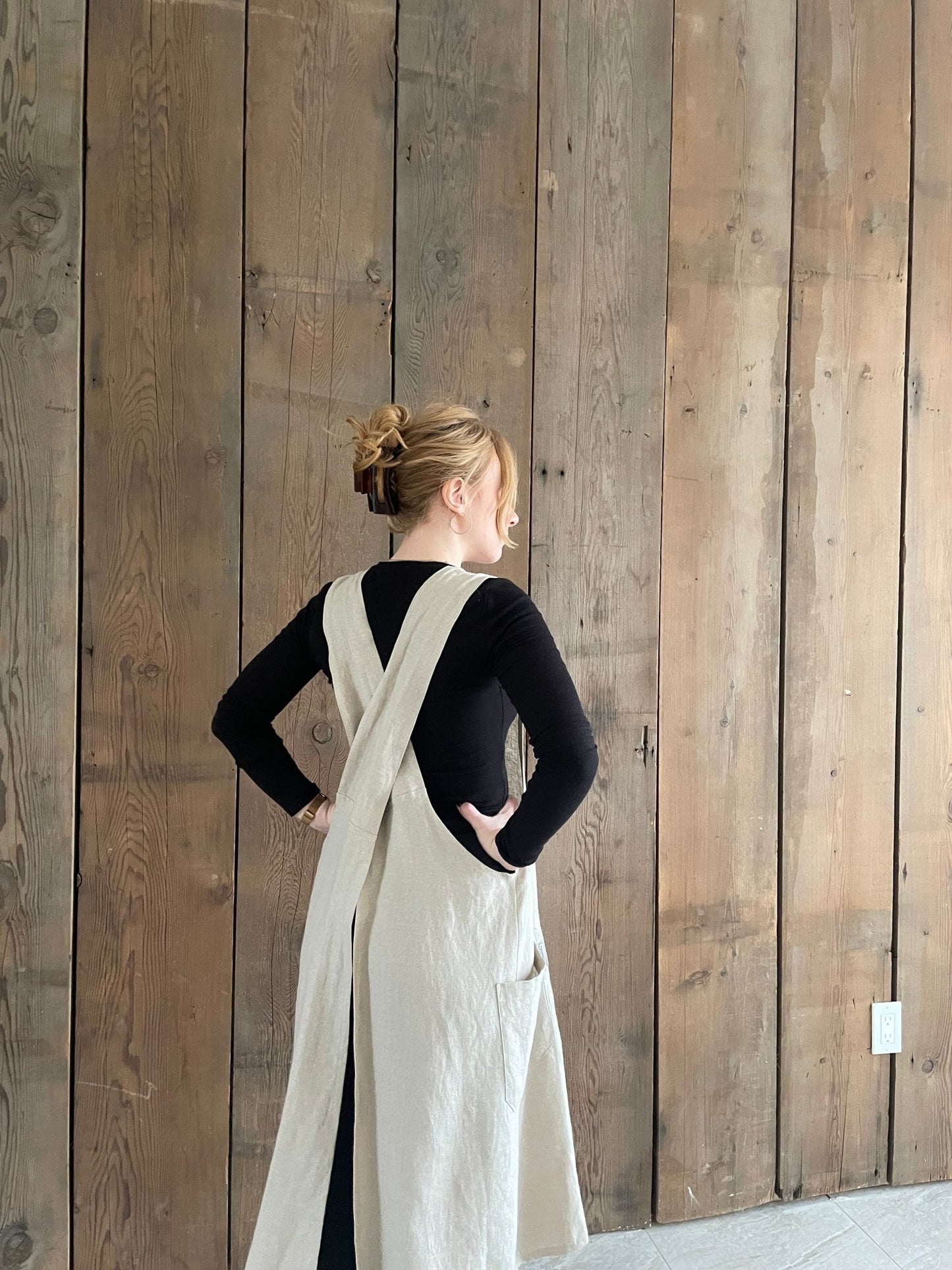 Rear view of a woman wearing a natural flax Linen Japanese cross back pinafore apron, standing against a wooden background.