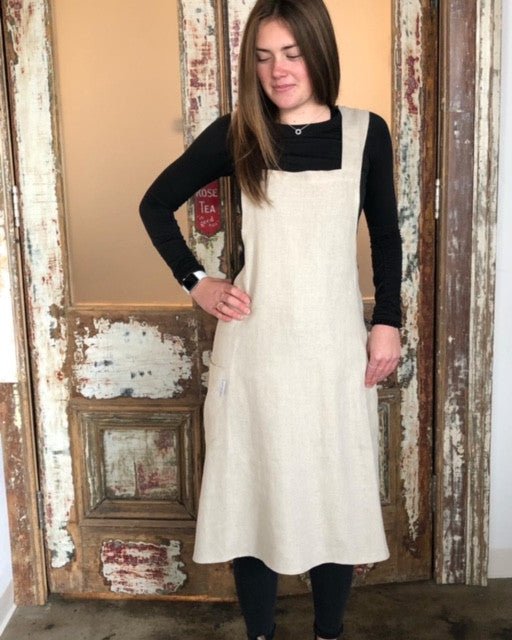 A woman wearing a natural flax Linen Japanese cross back pinafore apron, standing against an old distressed painted door.