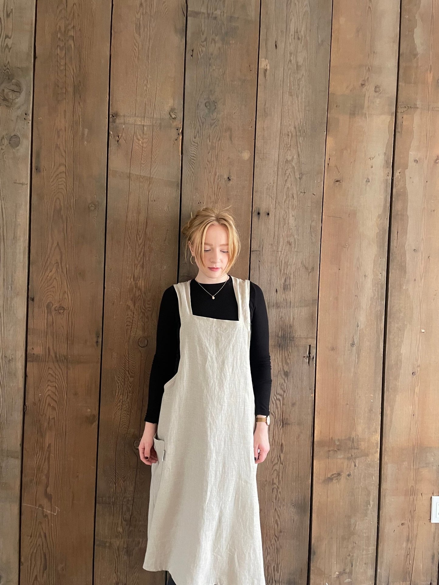 A woman wearing a natural flax Linen Japanese cross back pinafore apron, standing against a wooden background.