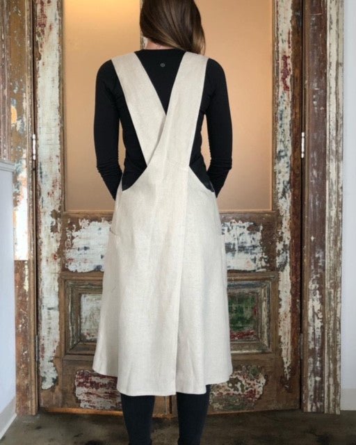 Rear view of a woman wearing a natural flax Linen Japanese cross back pinafore apron, standing against an old distressed painted door.