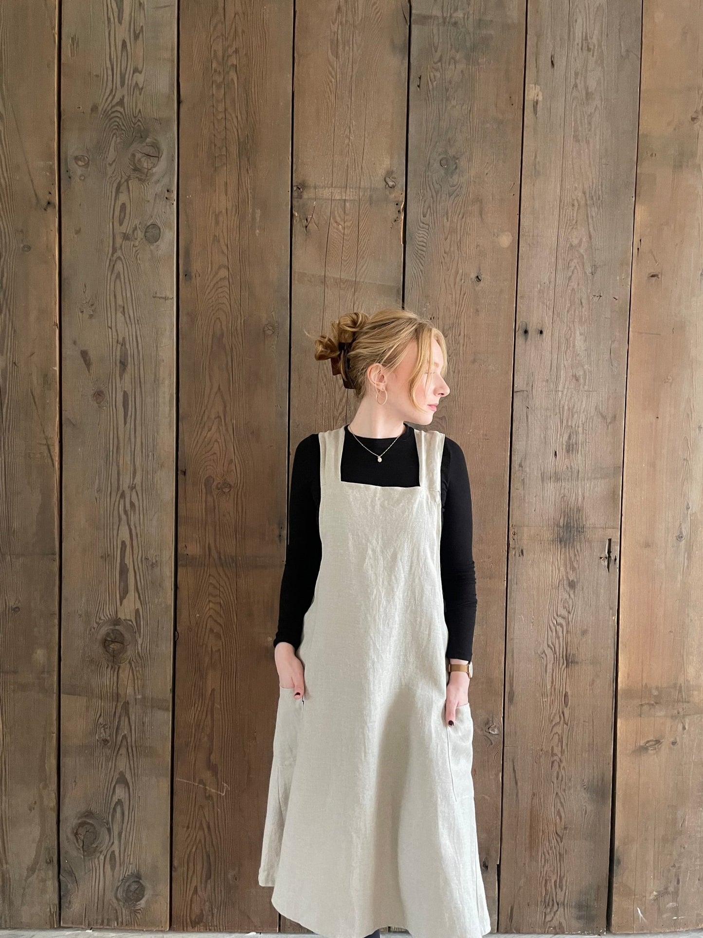 A woman wearing a natural flax Linen Japanese cross back pinafore apron, standing against a wooden background.