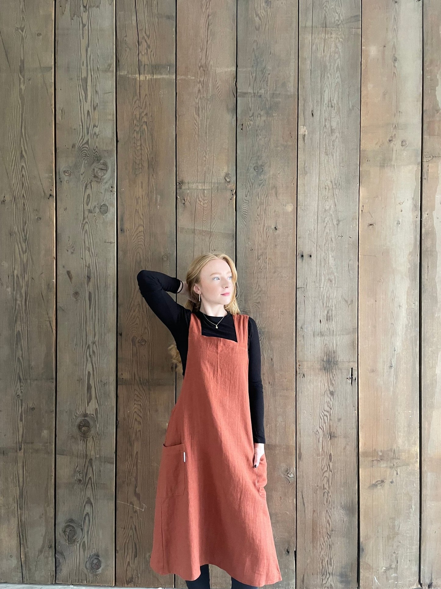 A woman wearing a rust copper coloured linen Japanese cross back pinafore apron, standing against a wooden wall.