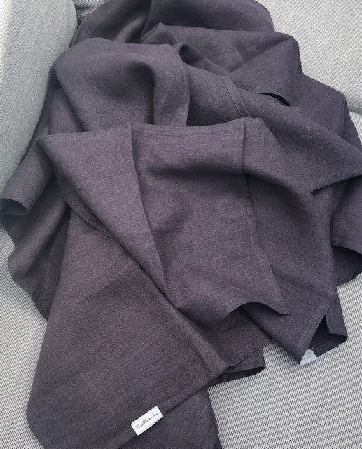Stonewashed European Oeko-Tex Certified Organic Linen Bath Towels in Dark Aubergine Purple with Rural Route Seven logo brand label and Simple Beauty, Every Day label, resting piled in gentle folds on a poolside sofa with beams of sunlight