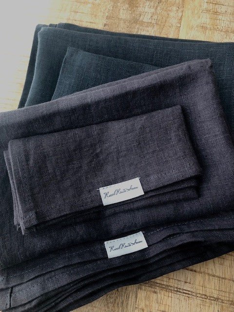 Asymmetrically placed stack of Stonewashed European Oeko-Tex Certified Organic Linen Bath Towels in Dark Aubergine Purple with Rural Route Seven logo brand label resting on top of dark charcoal grey stack of Stonewashed European Oeko-Tex Certified Organic Linen Bath Towels on an antique distressed pine table top with beams of sunlight