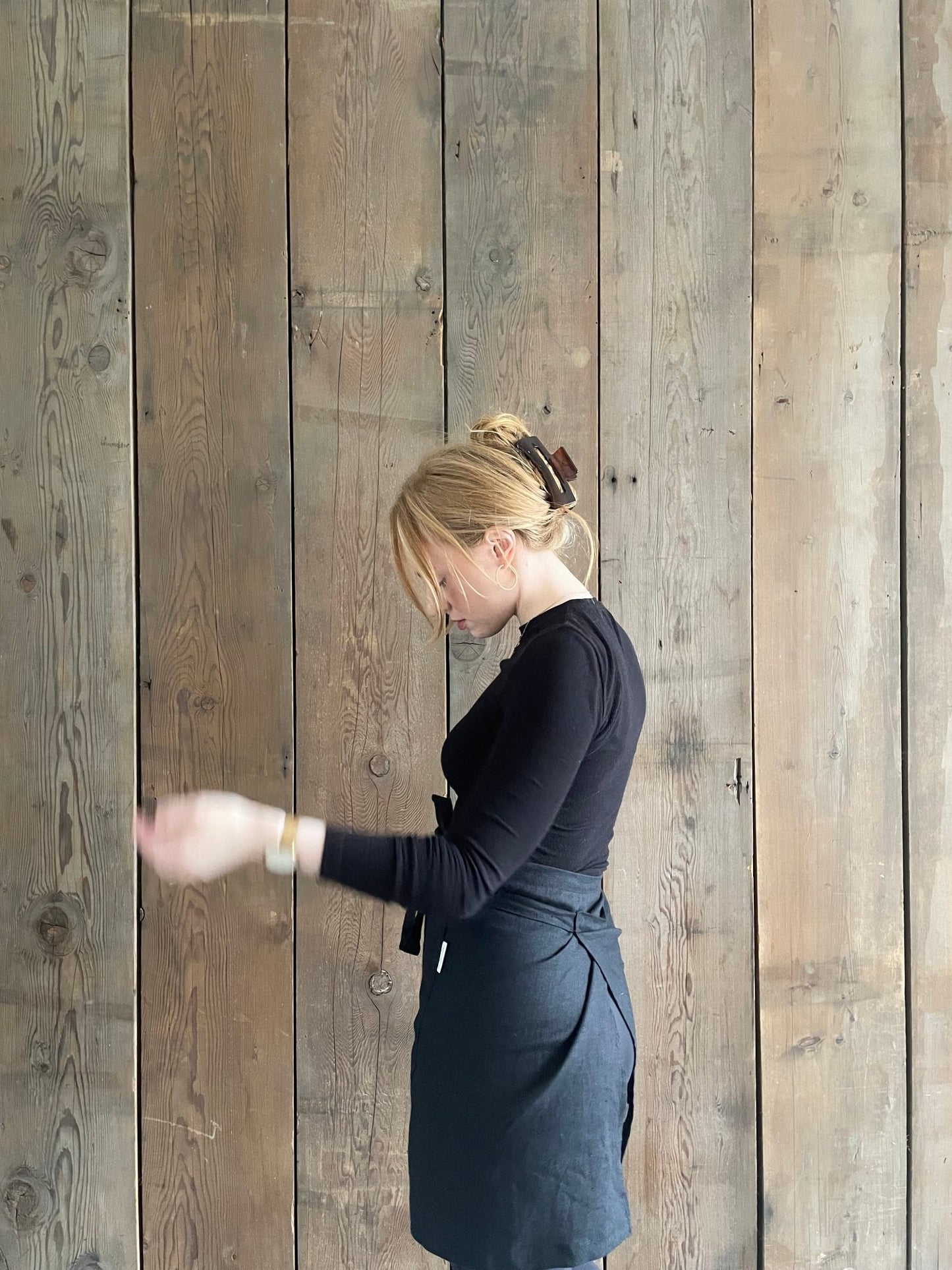 Side and rear view of young red haired woman wearing Black Oeko-Tex Certified Organic Linen Half Apron with Rural Route Seven logo brand label, black long sleeve t-shirt with black pants and rose gold necklace and small hoop earrings, hair up in clip with romantic wisps hanging down, standing against a rustic wood panelled wall