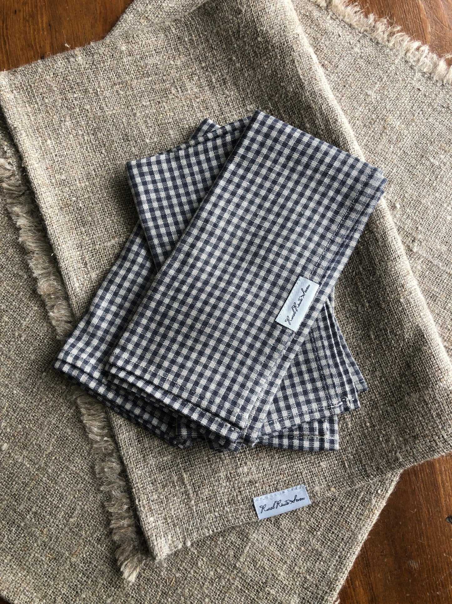 Four asymmetrically placed folded small charcoal grey and natural flax checks stonewashed European linen napkins with Rural Route Seven logo brand label, resting on one folded burlap linen natural flax placemat and one open burlap linen natural flax placemat with Rural Route Seven logo brand label, resting on distressed antique pine table top with beam of sunlight