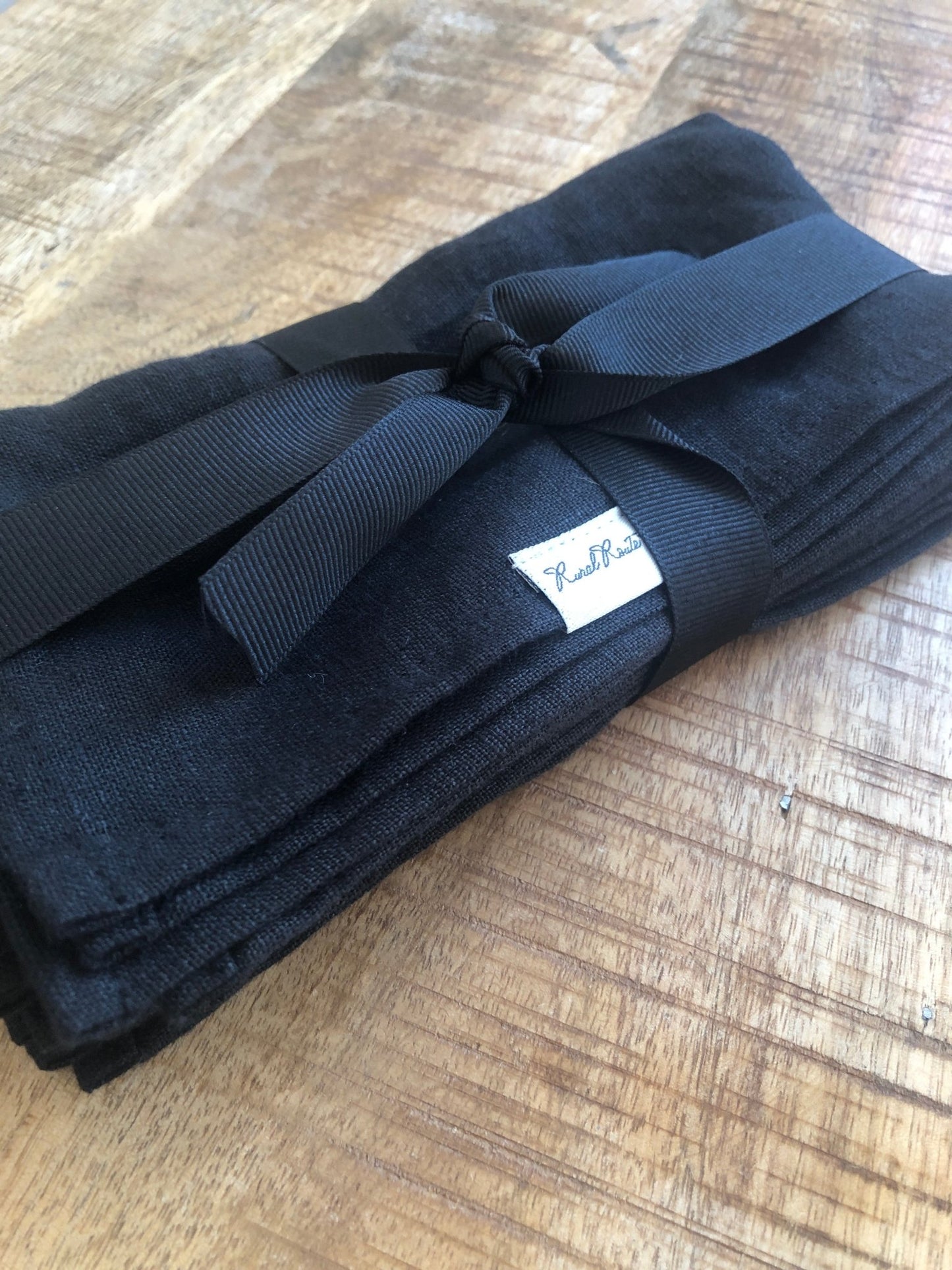 Close up of folded black stonewashed European linen napkins with Rural Route Seven logo brand label, wrapped and tied in black grosgrain ribbon, resting on distressed antique pine table top