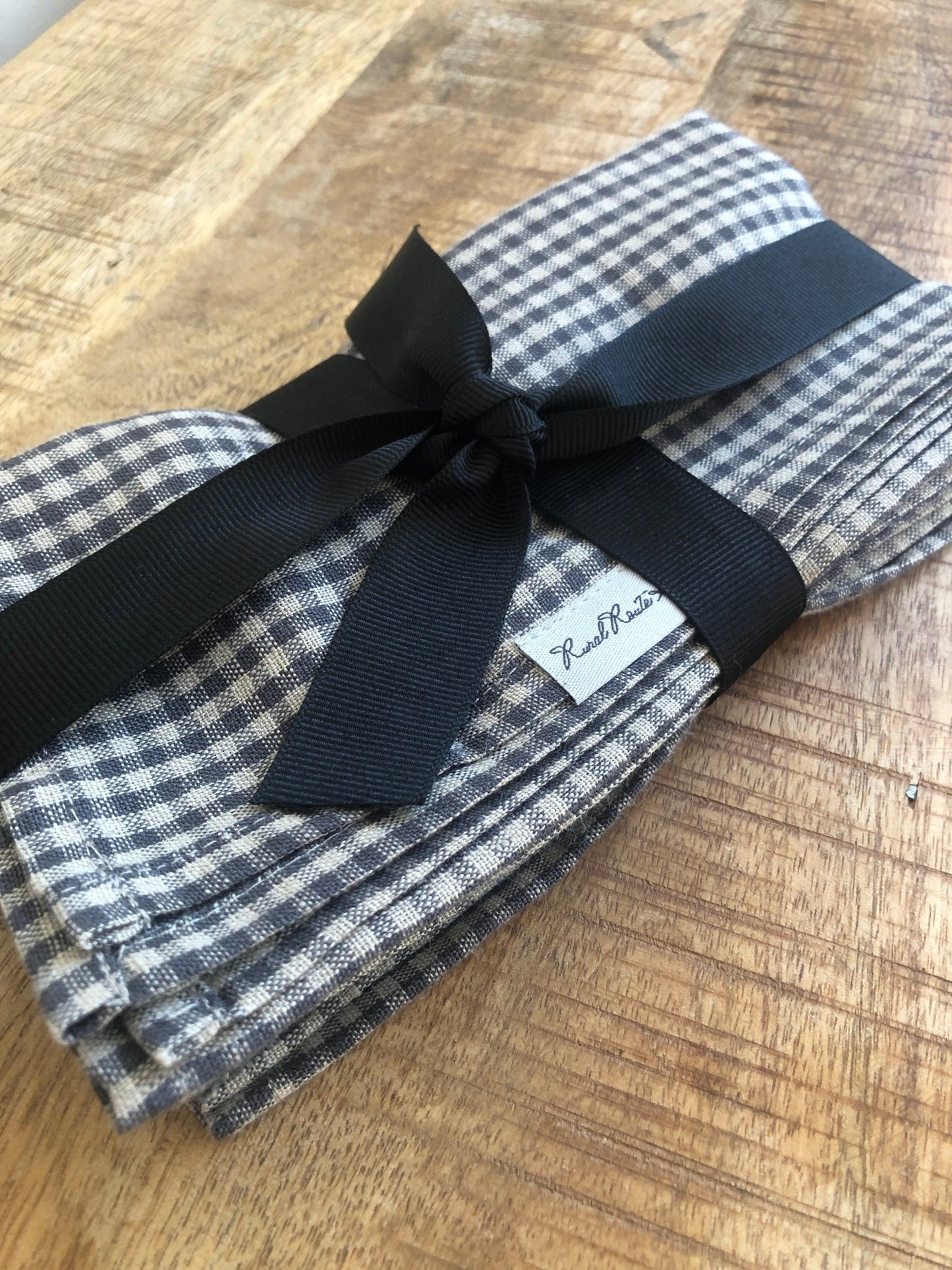 Close up of folded small charcoal grey and natural flax checks stonewashed European linen napkins with Rural Route Seven logo brand label, wrapped and tied in black grosgrain ribbon, resting on distressed antique pine table top