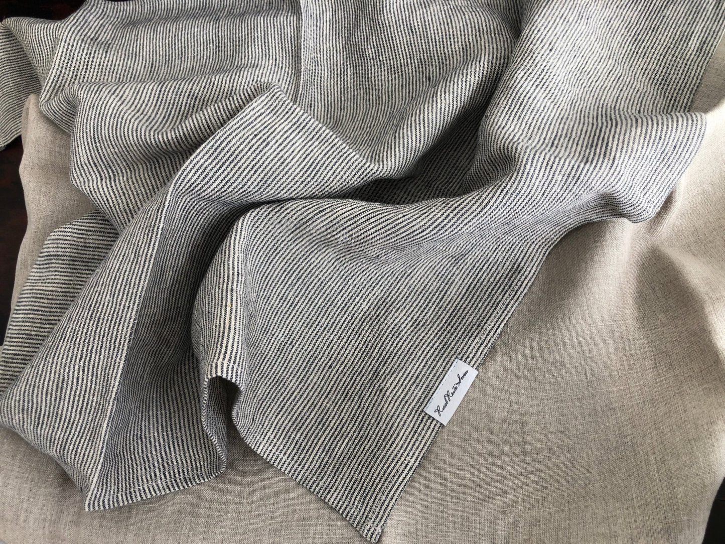 close up of open black and natural flax striped stonewashed European linen tea towel with Rural Route Seven logo brand label, and natural flax linen stonewashed European tea towel with gentle folds resting on distressed antique sideboard with beams of sunlight