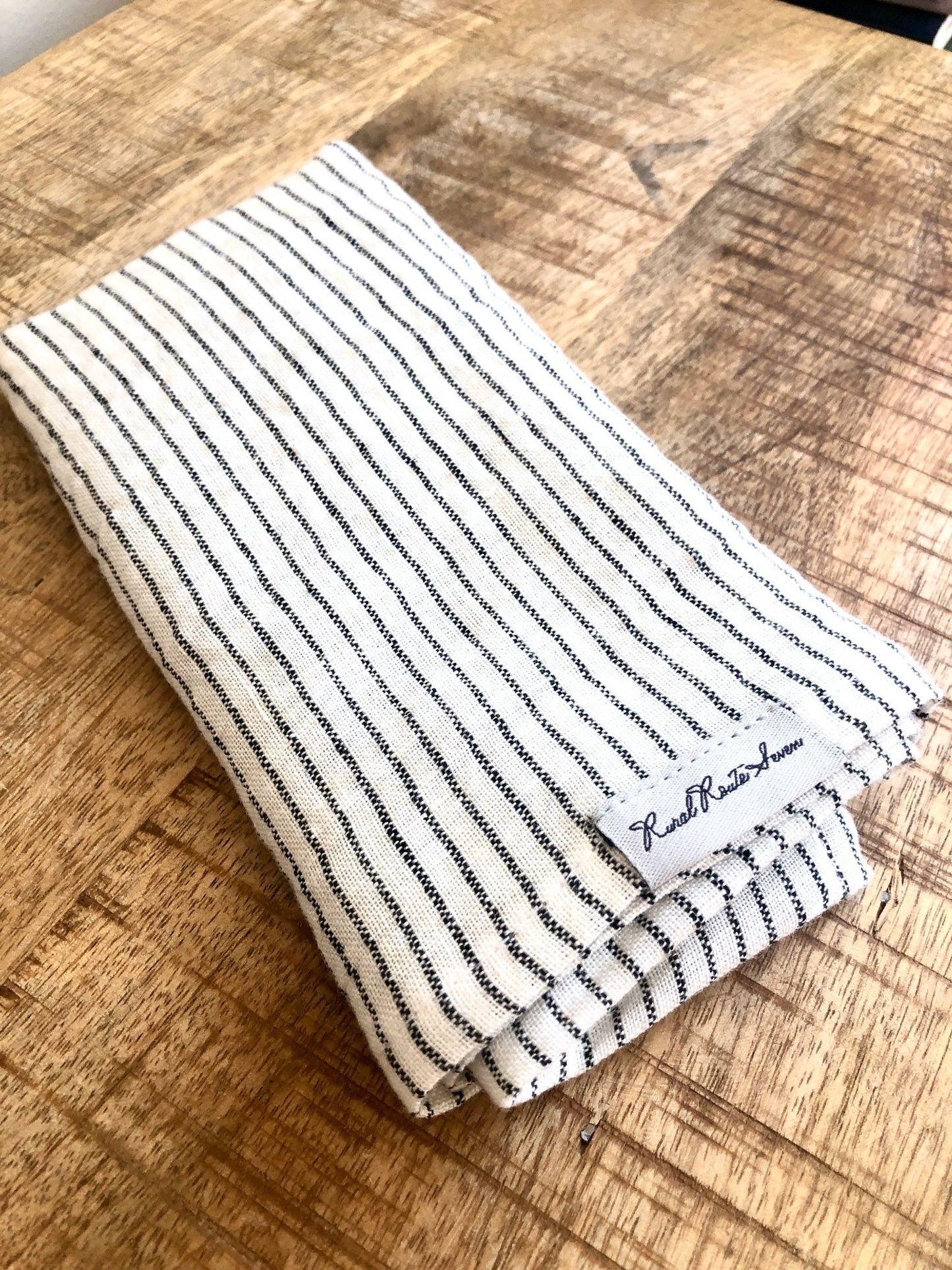close up of folded White with Black Stripes Stonewashed Linen Tea Towel with Rural Route Seven logo brand label, on distressed antique pine table