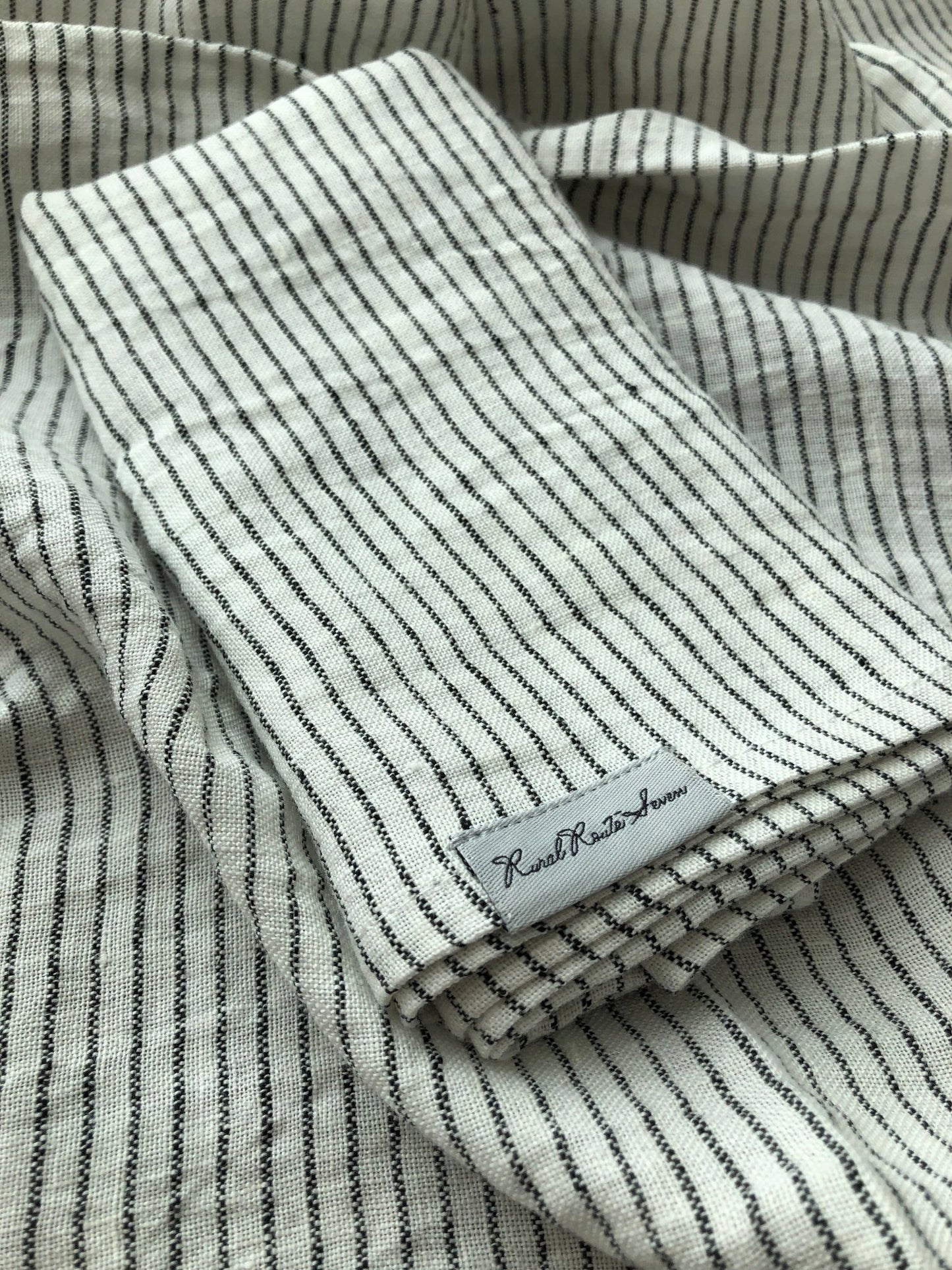 Close up of folded White and Black Stripes European Linen Tea Towel with Rural Route Seven logo brand label resting on an open tea towel of the same black and white striped fabric