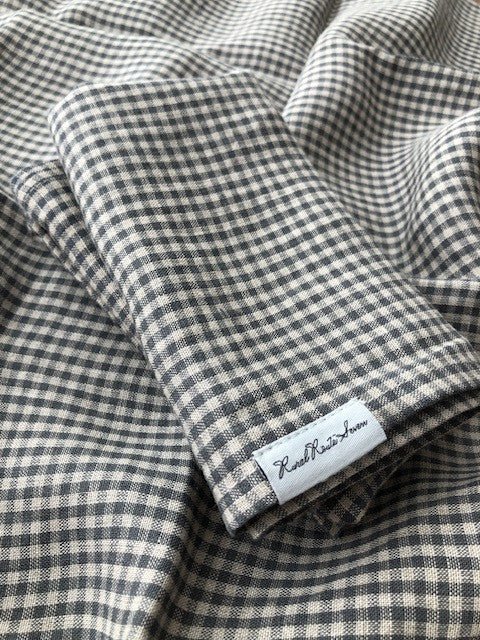 Folded natural and small grey checked linen tea towels with Rural Route Seven tag on lower corner lying on same open tea towels in background