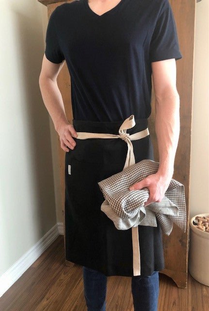 Young tall and thin caucasian male chef wearing denim Pullin' jeans, a navy V-neck t-shirt and a black linen apron with natural ties and holding stonewashed European Oeko-Tex Certified Organic Linen Tea Towels with Rural Route Seven logo brand label in small charcoal grey and natural flax checks, with antique pine cupboard and maple hardwood floors, and antique crock pot filled with wine corks in the background