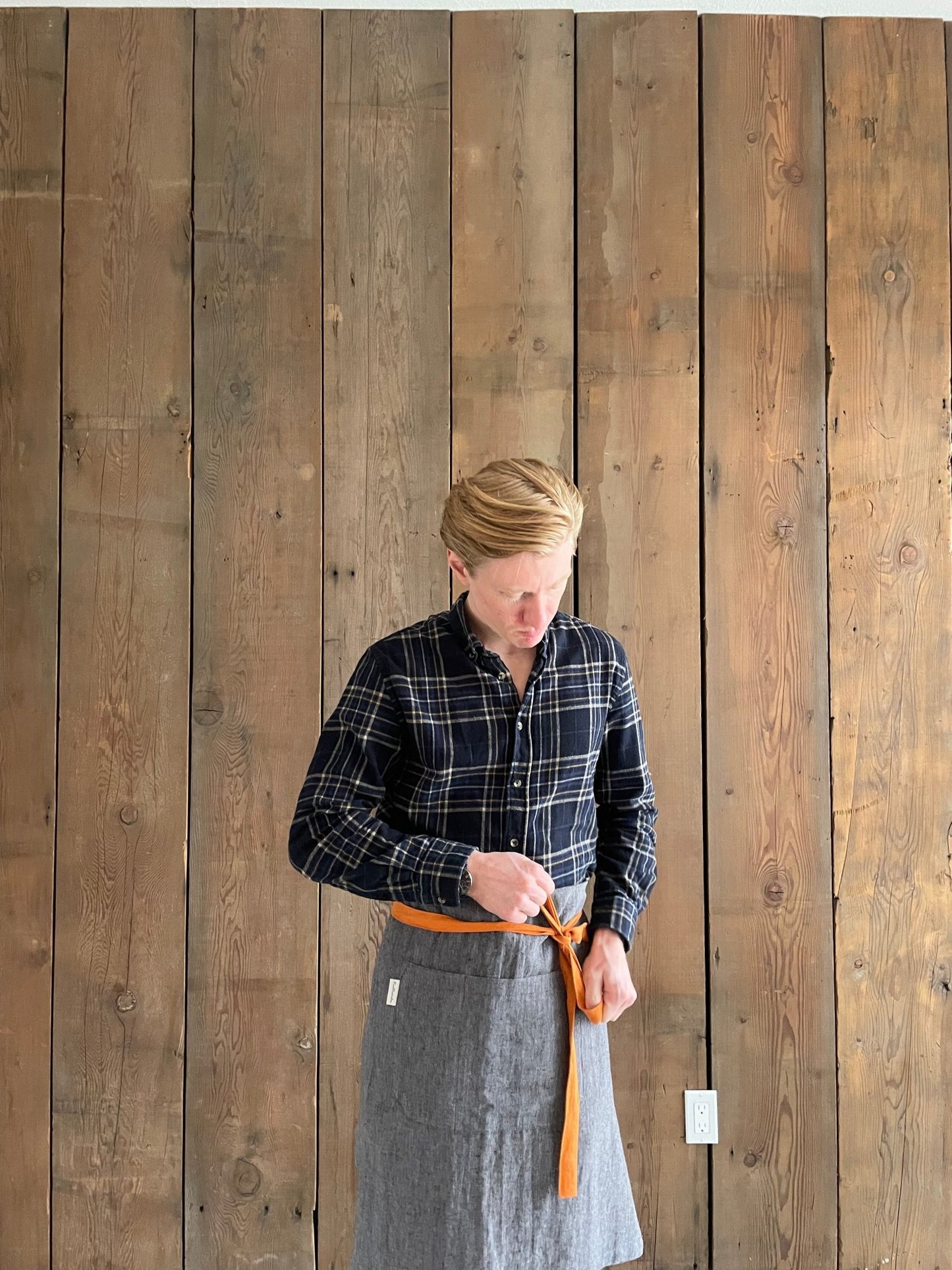 Young man wearing navy, gold and white flannel shirt and grey black melange stonewashed Oeko-Tex Certified Organic Linen Men's Bib Apron with pumpkin orange canvas ties and Rural Route Seven logo brand label, apron is folded into half apron showing pumpkin orange canvas ties; old wood panel wall in background