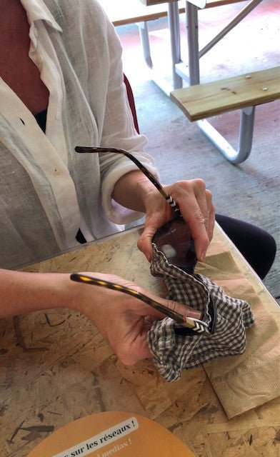 Torso of stylish Caucasian women wearing stonewashed European Linen blouse sitting at rustic chip board cafe table at Jean Talon Market in Montreal cleaning Burberry sunglasses with Organic Oeko-Tex Certified Stonewashed European Linen Handkerchief from Rural Route Seven brand. Picnic table and bright beams of sunlight in the background