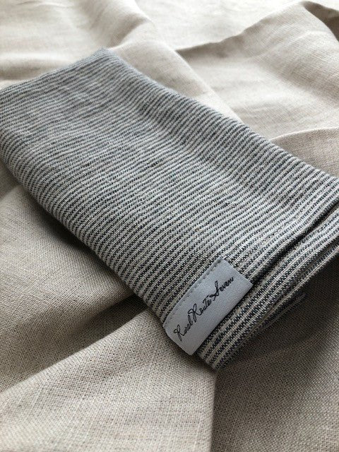 close up of folded black and natural flax linen striped stonewashed european linen tea towel with Rural Route Seven logo brand label, resting on open natural flax linen stonewashed linen tea towel with beams of sunlight