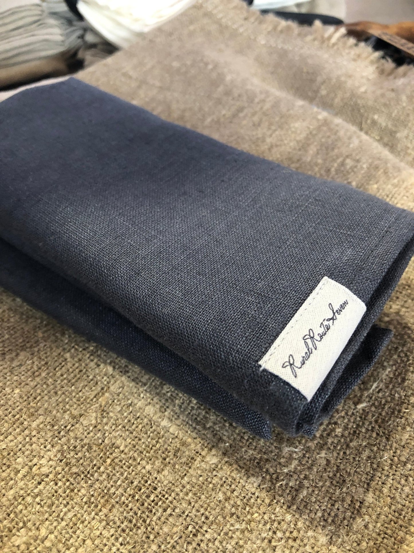 Close up of folded charcoal grey stonewashed European linen tea towel with Rural Route Seven logo brand label, resting on burlap natural flax placemat with beam of sunlight and a stack or pile folded tea towels in the background