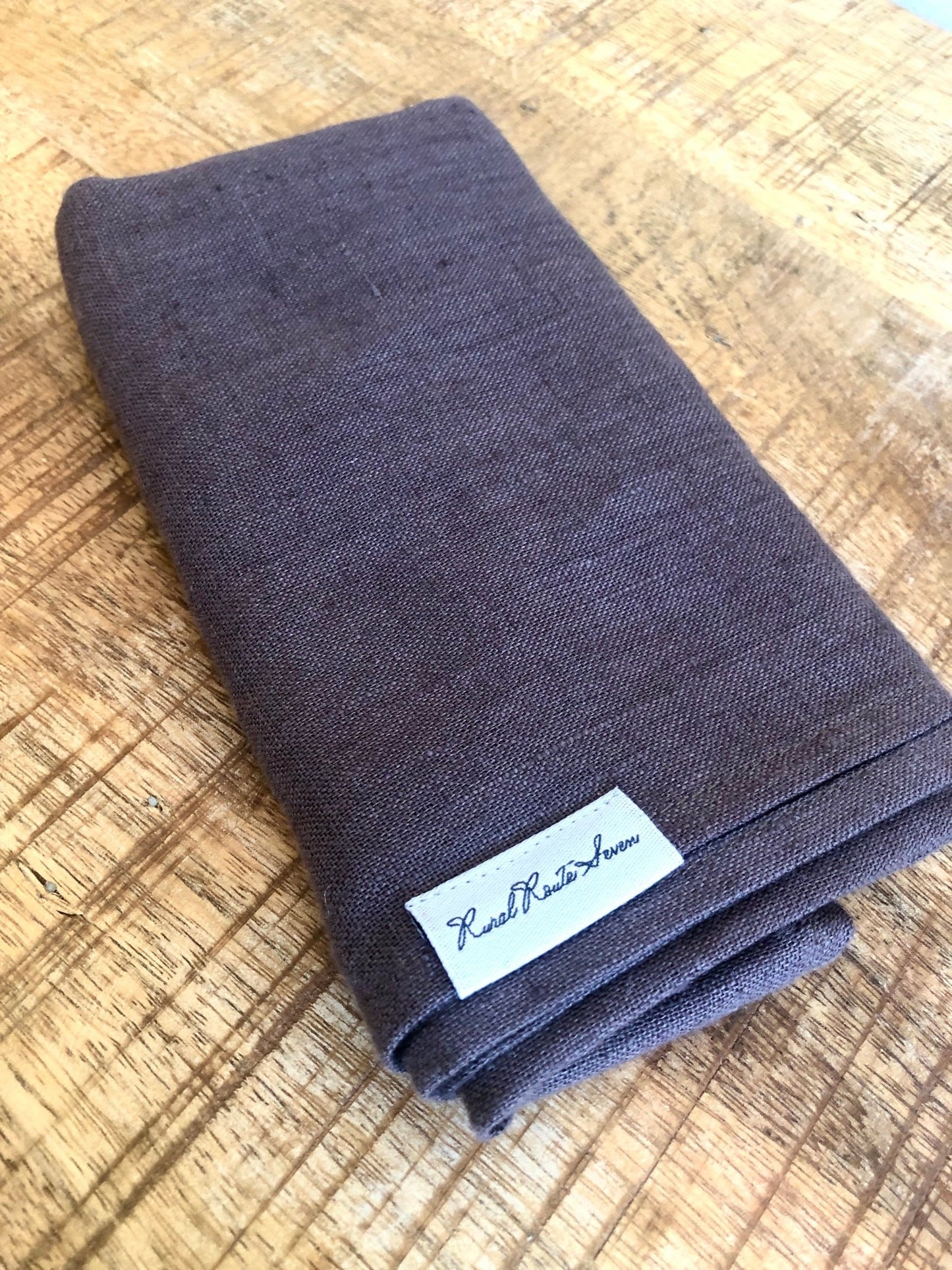 Close up of folded Dark Aubergine deep dark purple stonewashed European linen tea towel with Rural Route Seven logo brand label, resting on distressed antique pine table top