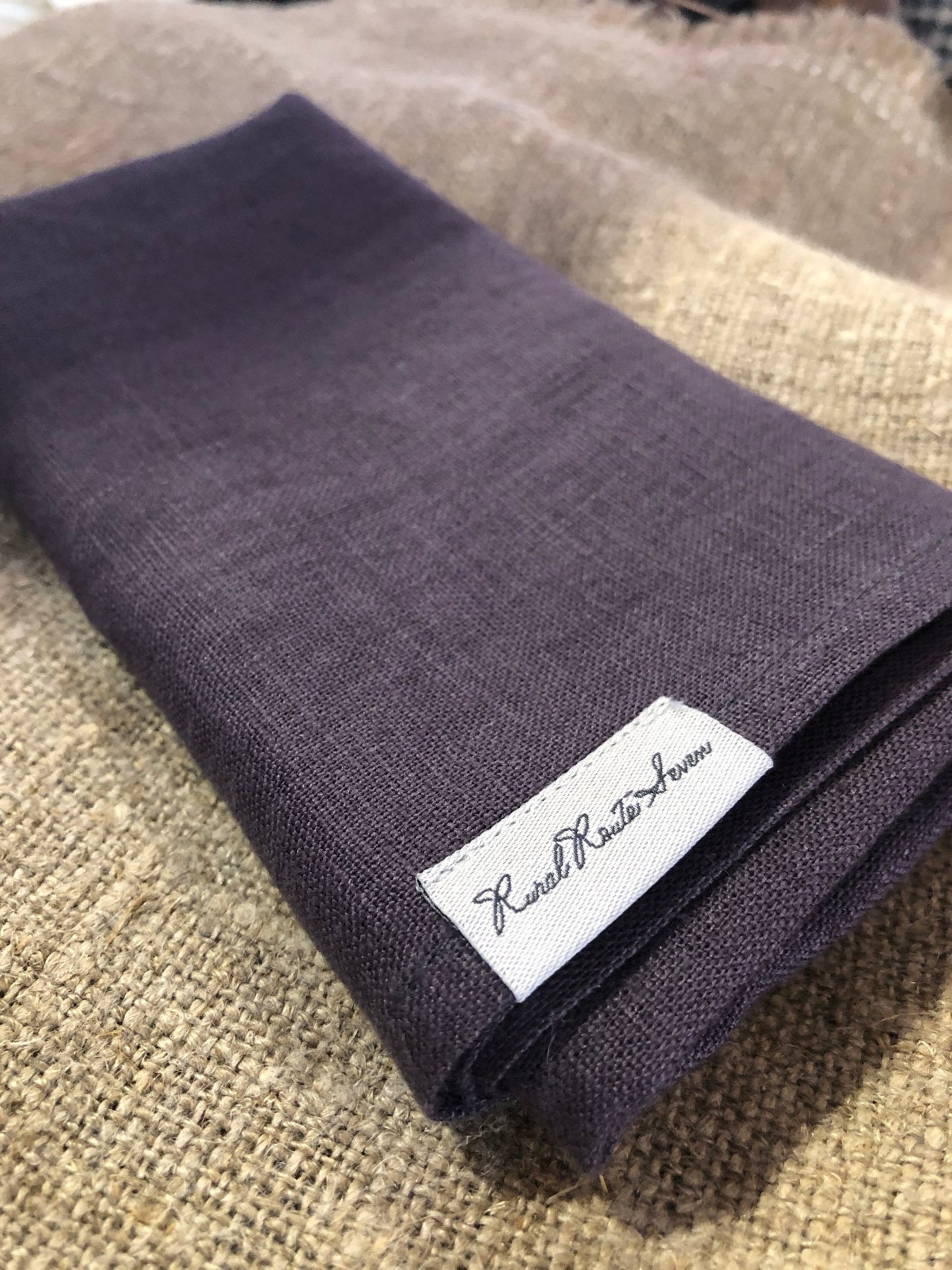 Close up of folded Dark Aubergine deep dark purple stonewashed European linen tea towel with Rural Route Seven logo brand label, resting on burlap natural flax linen placemat with beam of sunlight