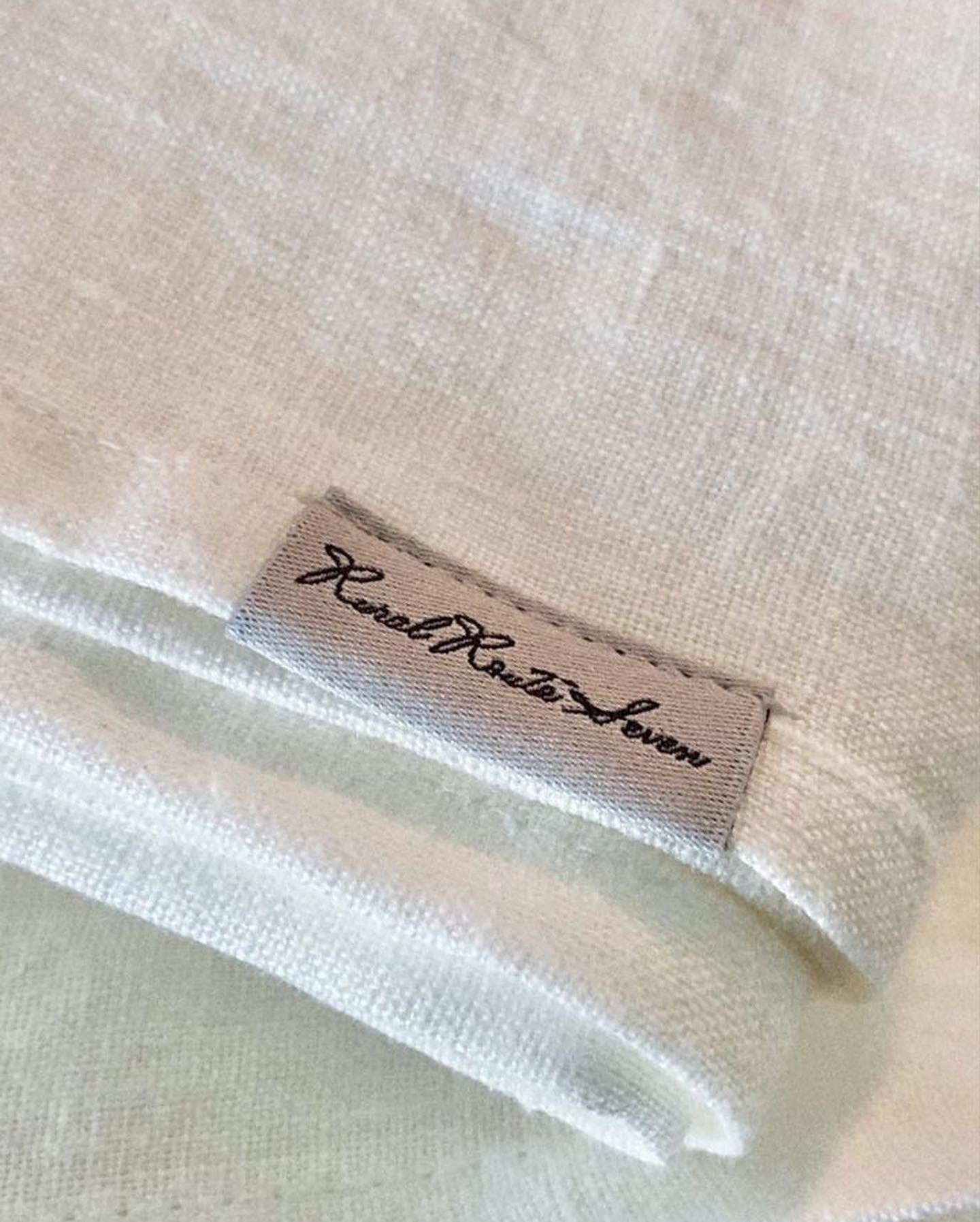 Close up of folded edges of off-white textured stonewashed European linen tablecloth with Rural Route Seven logo brand label with grey background and black font print