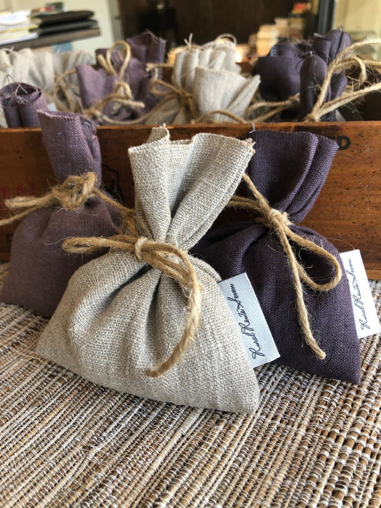Close up of Stonewashed European Linen and Canadian Lavender Sachets in natural flax, dark aubergine purple, dusty light lavender purple with Rural Route Seven logo brand label, tied with natural twine,  resting on textured natural placemat with honeyed antique rectangular cheese box in the background holding many stonewashed European linen and Canadian lavender sachets in natural linen, dark aubergine purple and dusty light lavender purple tied with natural twine