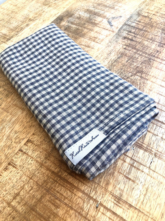 Folded natural and small grey checked linen tea towel with Rural Route Seven tag on lower corner against wood table top