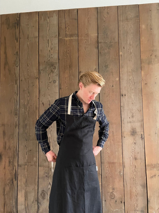 Man wearing black stonewashed Men's Linen Apron with natural canvas ties and Rural Route Seven label, tying the apron